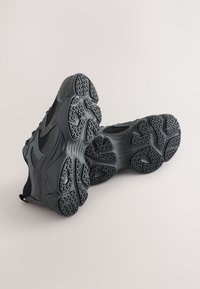 Baskets de sport en mesh noir et matériau synthétique, avec des semelles texturées dotées de crampons à motifs et un design élégant et aérodynamique.