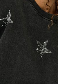 Sweat-shirt noir avec deux étoiles ornées de strass sur les épaules. Le tissu a une texture douce avec une légère brillance.