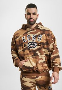 Brun camo hættetrøje med hvidt logo, kengurulomme, ribbede manchetter og kant. Matchende camo sweatpants fuldender outfittet.