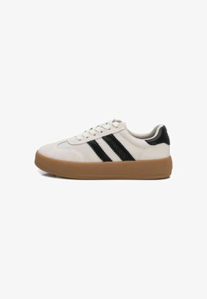 Zapatilla blanca con puntera de ante, rayas negras y acento negro en el talón. Presenta un diseño de cordones y una suela de goma para un mejor agarre.