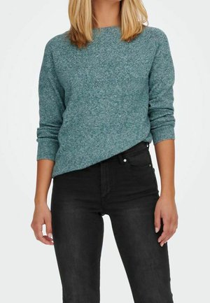 Pull en maille teal avec un col rond et des poignets côtelés, associé à un jean noir ajusté. Texture douce et coupe décontractée.