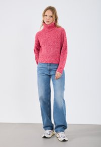 Maglione a collo alto rosso a maglia con un motivo testurizzato, abbinato a jeans a gamba larga blu chiaro e sneaker multicolori. Sfondo bianco uniforme.