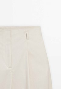 Beżowe spodnie o gładkiej fakturze, z plisami z przodu, płaskim pasem i bocznym detalem w okolicy talii.