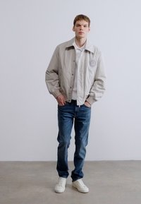 Svijetlosiva skraćena jakna s kopčanjem na gumbe, s okruglim zakrpom, uparena s plavim trapericama i bijelim tenisicama na neutralnoj pozadini.