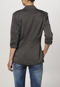 Grijze op maat gemaakte blazer met driekwartmouwen, gestructureerde achterkant en opgehoopte details op de mouwen, gecombineerd met blauwe denim jeans.