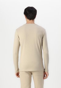 Camisa beige de manga larga con cuello redondo, que presenta una textura suave y un detalle de costura mínimo. Combinada con pantalones beige a juego.