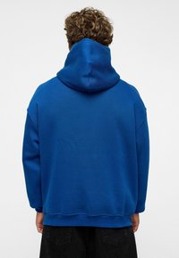 Blauwe hoodie met een relaxte pasvorm, voorzien van een grote capuchon en geribbelde manchetten. Gemaakt van een zachte stof, heeft het een gladde textuur.