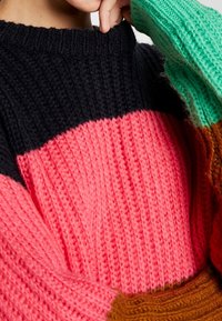 Pull en maille épaisse avec des sections multicolores : noir, rose, sarcelle et marron. Texture côtelée avec un col haut et des manches larges.