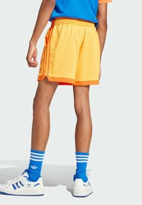 Bright yellow mesh shorts with an orange trim, side pockets, and three vertical stripes on the sides. Worn with blue socks and white shoes.