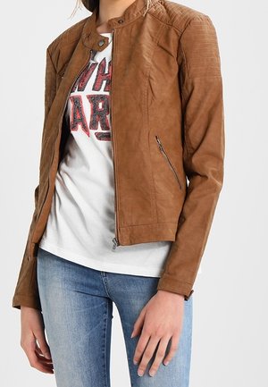 Personne portant une veste en cuir marron zippée sur un T-shirt blanc à motif graphique et un jean bleu clair, debout devant un fond uni.