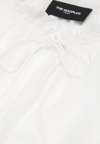 Vit blus med en mjuk, sidenliknande textur, som har en rund halsringning, knytning fram och veckade detaljer längs framsidan. Varumärkesetikett synlig.