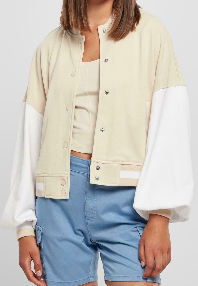 Béžová a bílá zkrácená bunda s knoflíkovým zapínáním, oversized rukávy a pásem na spodním lemu, doplněná o světle modré šortky. Hladká textura.