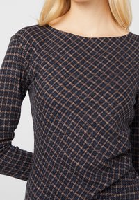 Donna che indossa un top aderente a maniche lunghe con un motivo a scacchi in blu navy e marrone e un collo ampio e arrotondato.