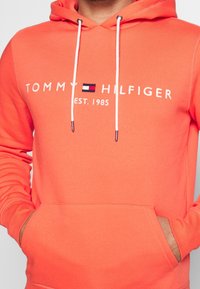 Felpa arancione con tasca frontale, cordini bianchi e testo ricamato "TOMMY HILFIGER EST. 1985," con un piccolo motivo a blocchi di logo.