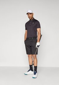 Svart pikétröja med en logotyp, svarta shorts, vita golfskor med blå detaljer, håller en golfklubba och bär en vit keps och handske.