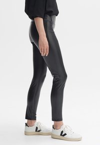 Svarta leggings i konstläder med hög midja, slät textur och figurnära design, tillsammans med vita sneakers med svarta detaljer.