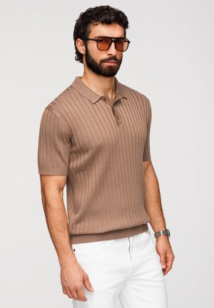 Mężczyzna z brodą w brązowej prążkowanej koszulce polo z krótkim rękawem, pomarańczowych okularach przeciwsłonecznych, białych spodniach i srebrnym zegarku na nadgarstku, stojący na białym tle.