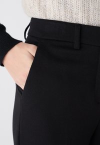Pantalons noirs ajustés avec une ceinture plate et une poche latérale. La texture est lisse, fabriquée à partir d'un tissu structuré.