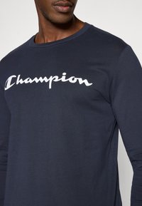 Maglietta a maniche lunghe blu marina in cotone con scollo rotondo, con logo bianco "Champion" in carattere corsivo sul petto.