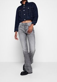 Chaqueta corta de mezclilla azul con cierre de botones, dos bolsillos en el pecho y cuello levantado. Pantalones vaqueros grises acampanados. Botas negras de plataforma.