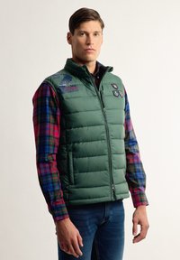 El chaleco acolchado verde presenta un cuello alto, cierre frontal y parches de logo. Se lleva sobre una camisa de cuadros multicolor de manga larga.