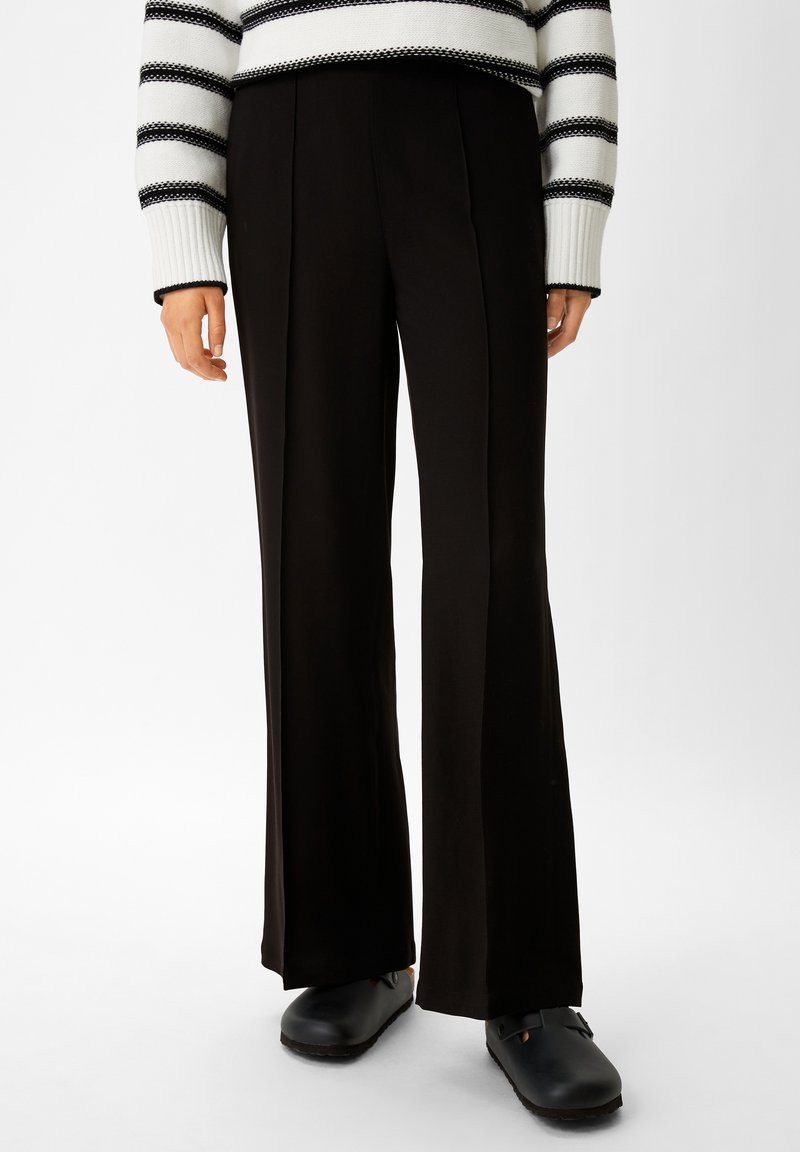 Black wide-leg trousers with a smooth fabric, featuring pleats along the front and a tailored fit, paired with black clogs.