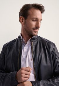 Czarna skórzana kurtka z wysokim kołnierzem, z przodu na zamek błyskawiczny, o subtelnym połysku, noszona na jasnoróżowej i białej koszuli w kratkę.