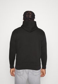Felpa nera con cappuccio, dal taglio morbido, realizzata in tessuto morbido, con tasca a marsupio e polsini a coste, indossata sopra jeans grigio chiaro.