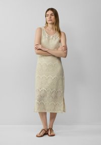 Strickkleid ohne Ärmel in Hellbeige mit spitzenähnlichen Mustern, gerader Silhouette und seitlichen Schlitzen, kombiniert mit braunen flachen Sandalen.