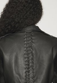 Personne aux cheveux bouclés portant une veste en cuir noire avec un laçage croisé qui descend verticalement dans le dos.