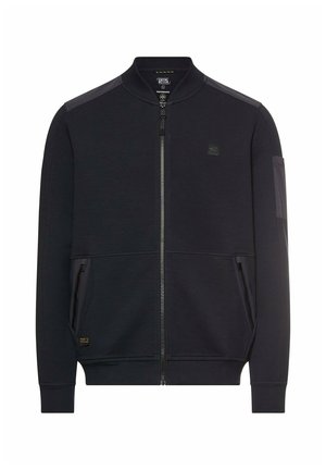 Sort bomberjakke med lynlås, ribstrikket krave og manchetter, to lynlåslommer foran samt et lille patch-logo på brystet og lomme på ærmet.