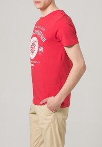 Rotes Baumwoll-T-Shirt mit kurzen Ärmeln, das einen weißen Grafikdruck und Beschriftungen auf der Vorderseite aufweist. Getragen mit beigen Hosen.