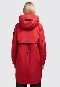 Chaqueta impermeable roja con cintura ajustable mediante cordón, capucha grande y puños con botones a presión. El material parece suave y brillante, con un logotipo en la parte trasera.