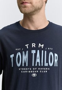 Camiseta de algodón azul marino con un estampado gráfico en azul claro y texto en blanco que dice "TOM TAILOR" con un estilo vintage en la parte frontal.