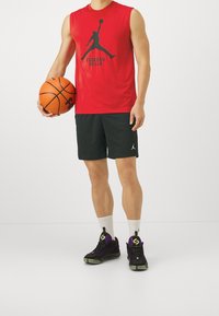 Rød ærmeløs trøje med en sort grafik, sorte shorts og sorte og lilla basketball-sko. Holder en basketball. Hvid baggrund.