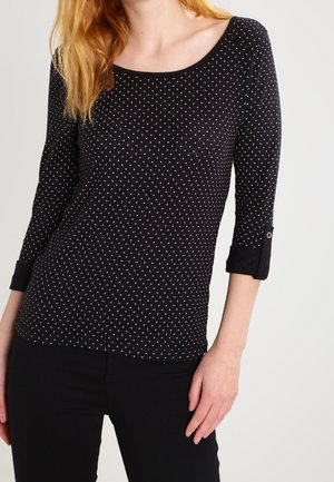 Femme portant un haut à manches longues noir à pois blancs et un pantalon noir, debout devant un fond clair uni.