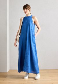 Blå linne maxi-klänning med rund halsringning, ärmlös design och sidfickor; kombinerad med vita sneakers. Slät textur och avslappnad passform.