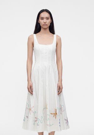 Femme aux longs cheveux foncés portant une robe blanche sans manches avec une broderie florale colorée près de l'ourlet, debout devant un fond blanc uni.