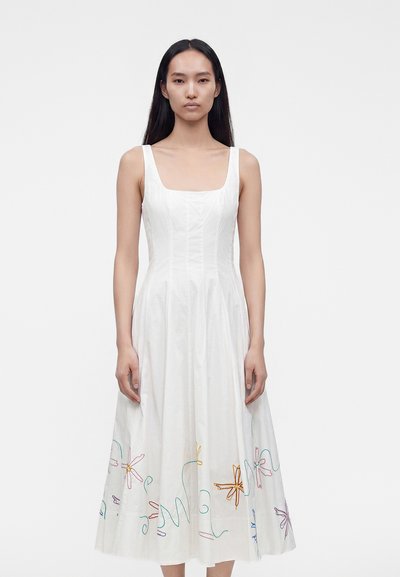 Femme aux longs cheveux foncés portant une robe blanche sans manches avec une broderie florale colorée près de l'ourlet, debout devant un fond blanc uni.