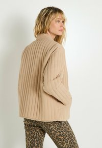 Femme blonde de profil portant une veste beige côtelée et un pantalon imprimé léopard, sur un fond clair uni.