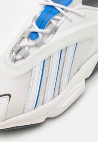 White and gray athletic shoe with blue accents, mesh upper, and thick rubber sole. Features intricate lacing and three white stripes on the side.