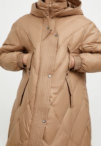 Beige gewatteerde jas met een hoge kraag en capuchon, voorzien van diagonale gestikte naden, drukknoopsluitingen en zijzakken. Gladde textuur.