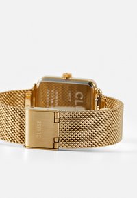 Montre avec un bracelet en mailles dorées et un boîtier rectangulaire. Le dos présente le nom de la marque gravé. Texture métallique lisse avec une finition brillante.