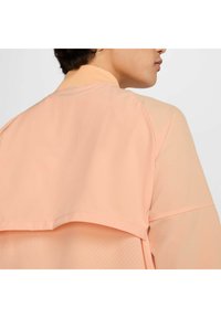 Chaqueta ligera de color melocotón con detalles de panel de malla, que presenta un escote redondo y una tela suave con silueta relajada.