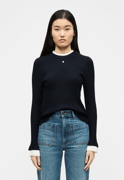 Νεαρή γυναίκα με μακριά σκούρα μαλλιά, φοράει μαύρο ριμπ πλεκτό πουλόβερ πάνω από λευκό τοπ και ψηλόμεσα μπλε τζιν με μεγάλες μπροστινές τσέπες.