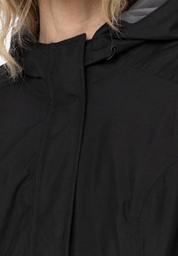 Svart vattentät jacka med hög krage, dragsko i huvan och synliga sömnadsdetaljer. Slät textur och funktionell design.