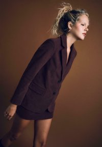 Ruskea corduroy-takki, jossa on kaksi nappia ja rintatasku, yhdistettynä yhdisteestä valmistettuun mini-hameeseen. Joustava tekstuuri, istuva muotoilu, täyspitkät hihat.