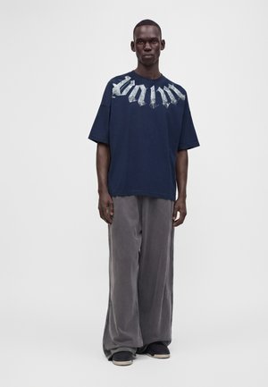 Personne grande à la peau foncée portant un t-shirt oversize graphique bleu marine et un pantalon ample en velours gris, debout sur un fond clair uni.