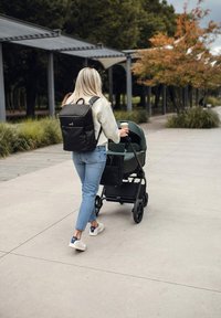 Černý batoh, džínové kalhoty a bílé tenisky. Osoba tlačí zelený kočárek s černou úložnou taškou, kráčí po dlážděné cestě pod stavbami.