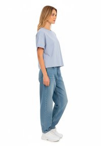 Sieviete stāv izsidusies, ģērbusies gaiši zilos oversized T-kreklos, platās zilās biksēs un baltajās sporta apavās uz baltas fona.